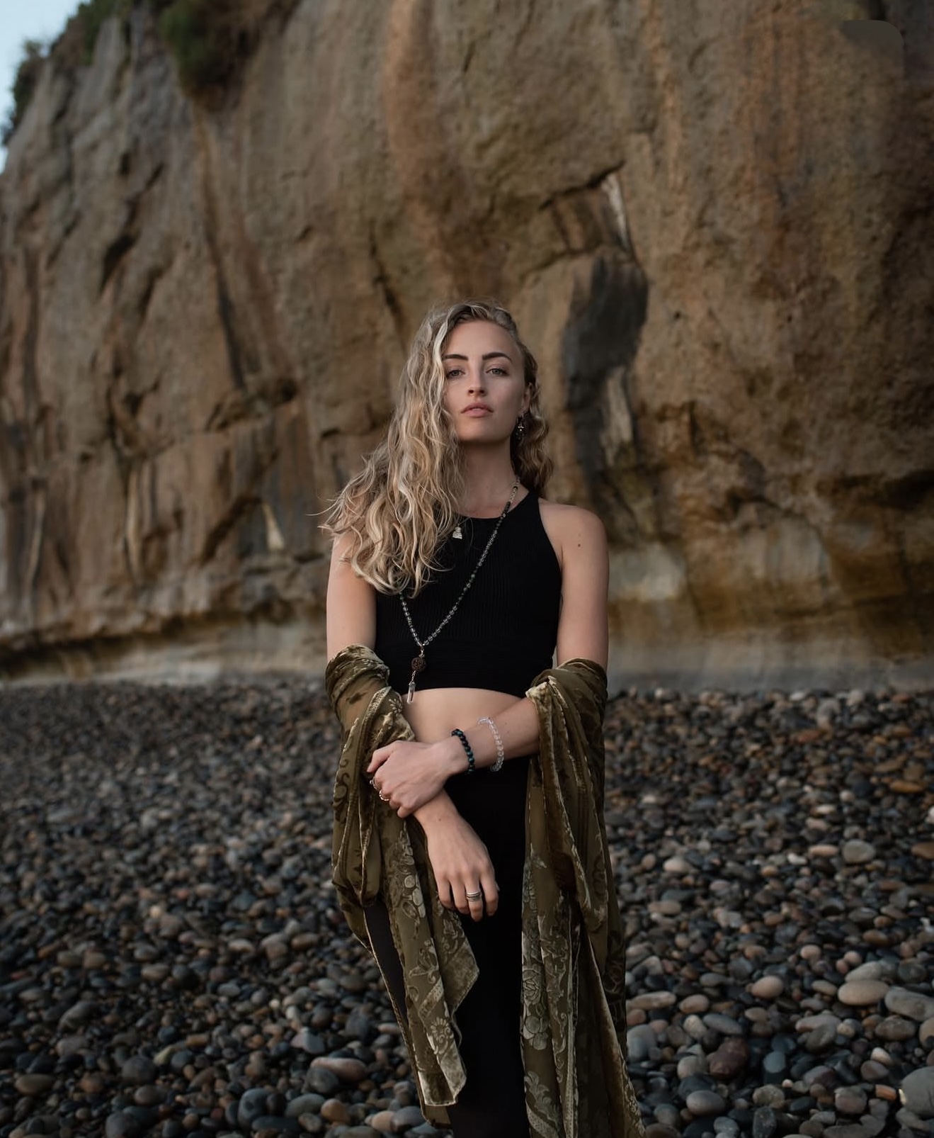 Jade Michael on a pebble beach in front of a sandstone cliff, an olive velvet shawl over her arms.